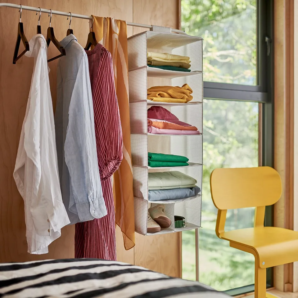 Bedroom storage: hanging clothes organiser, shelves with folded clothes, and chair by window.