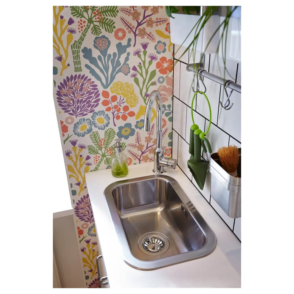 Stainless steel sink with tap and green soap dispenser. Colourful floral wallpaper behind.