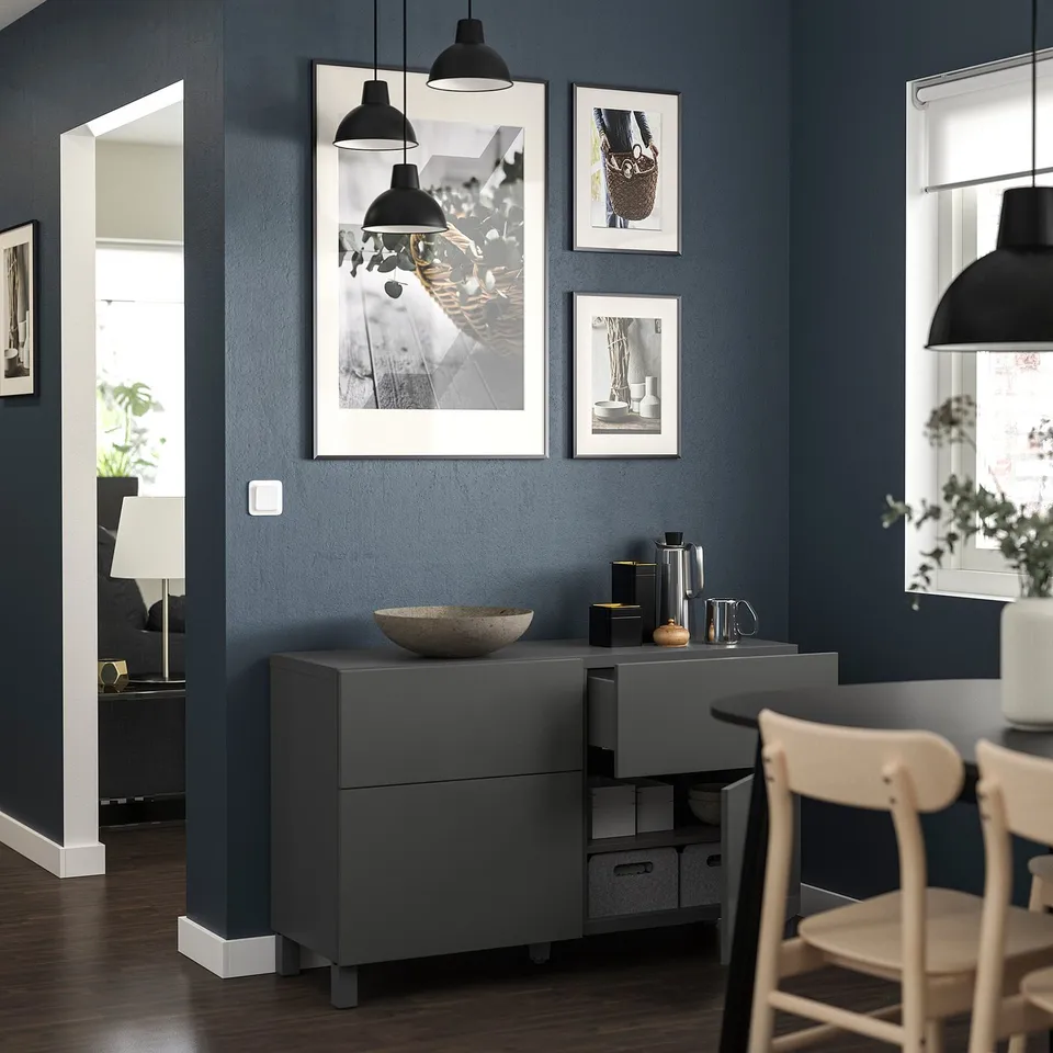 Modern living room with dark blue walls, black pendant lamps, framed art, grey storage unit, light wood chairs, and dark wooden floor.