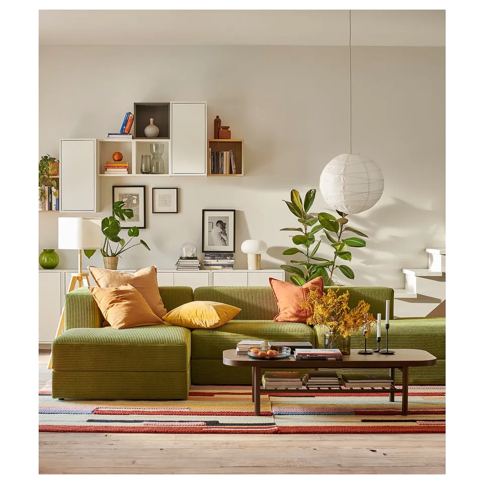Bright living room with green JÄTTEBO modular sofa, orange pillows, wooden coffee table, plants, colourful rug, and wall shelves with decor.