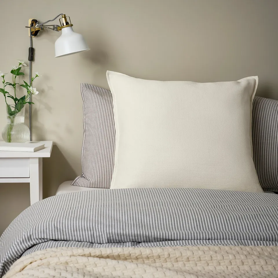 White LAGERPOPPEL cushion on striped bed, next to wall lamp.