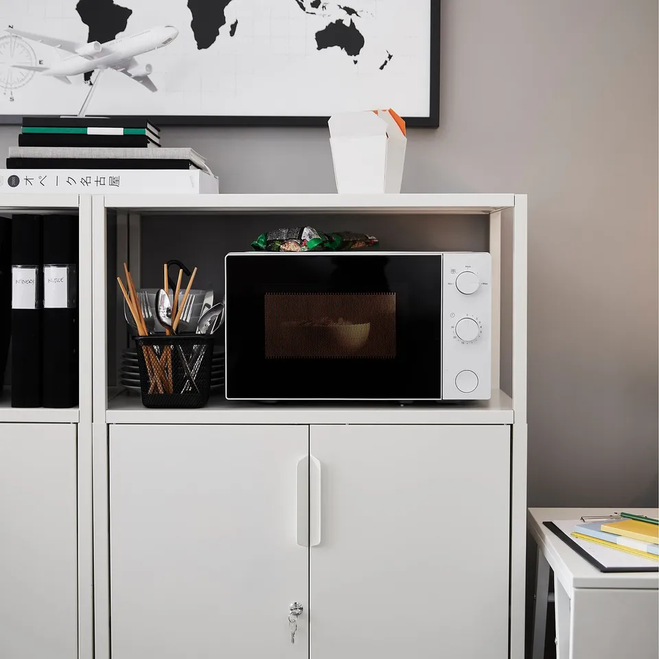 Small white TROTTEN microwave on office shelves. Next to utensils, books, and a world map. Fits neatly in compact space.