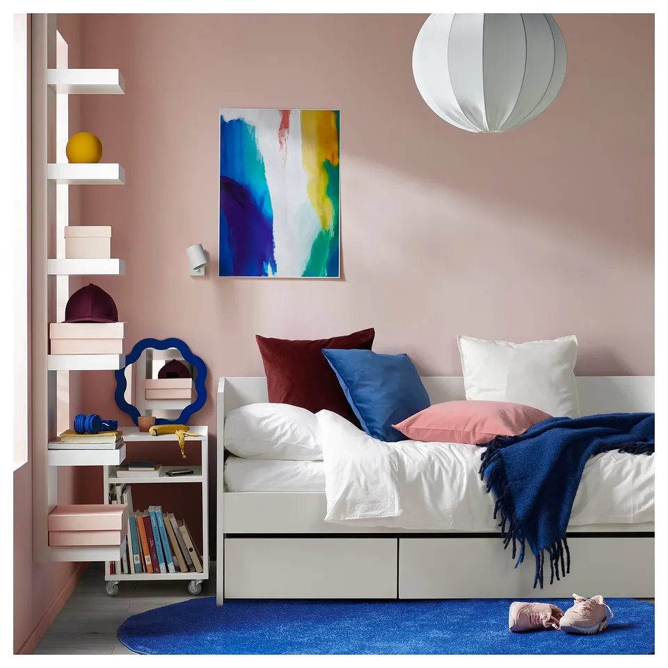 Modern bedroom with a white VIHALS daybed, colourful pillows, blue rug, abstract art, pink shelves with books, and a unique lamp.