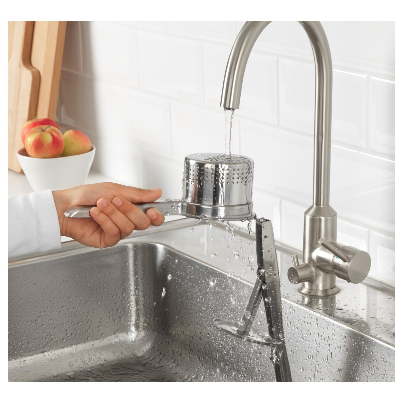 Stainless-steel kitchen sink with hands, water flowing, apples in background.