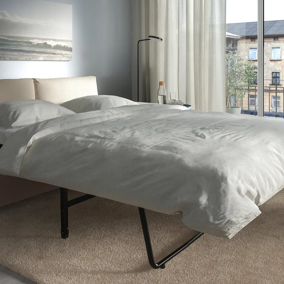 Modern bedroom with beige headboard, grey bedding, black metal bed. Tall lamp, bottles, sofa-bed option, city view.