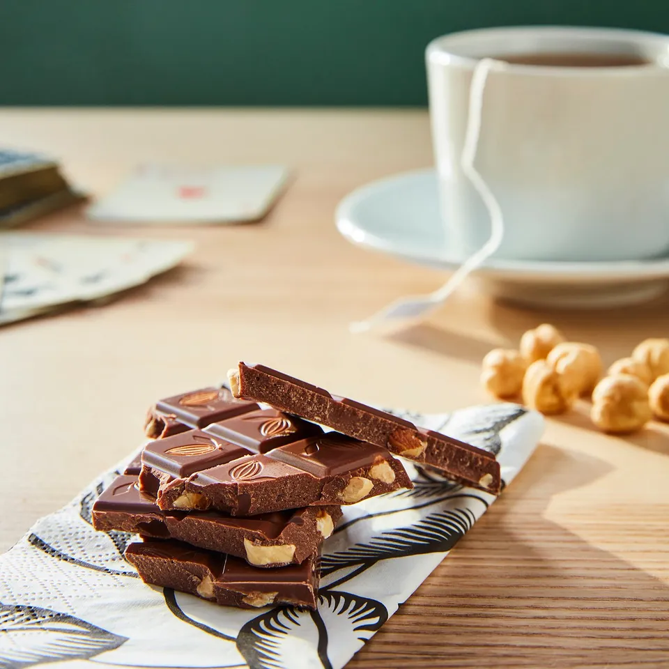 Stacked chocolate bars, some with nuts, on a patterned napkin near a coffee cup.