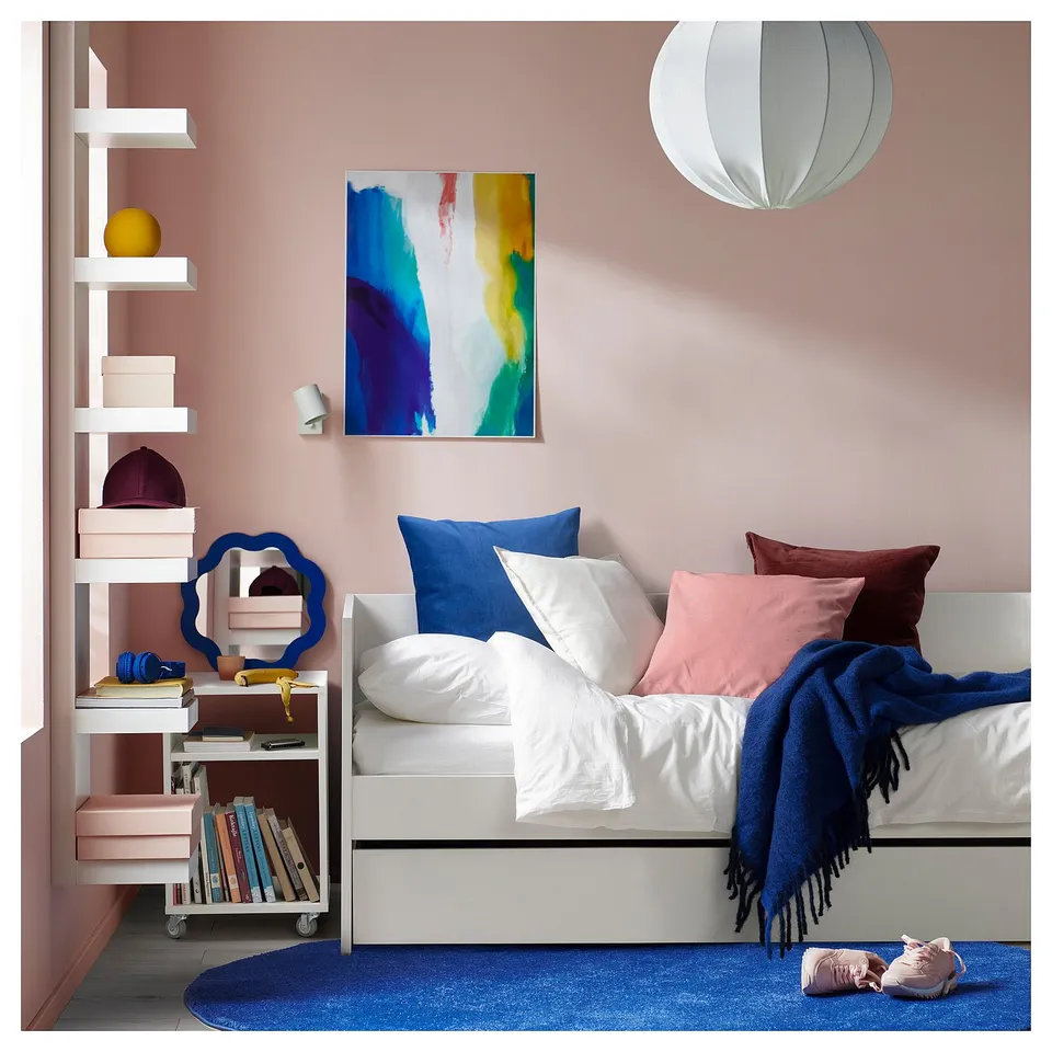 Modern bedroom with pink walls, white day-bed, blue rug, colourful pillows, abstract art, and white shelves with decorative items.