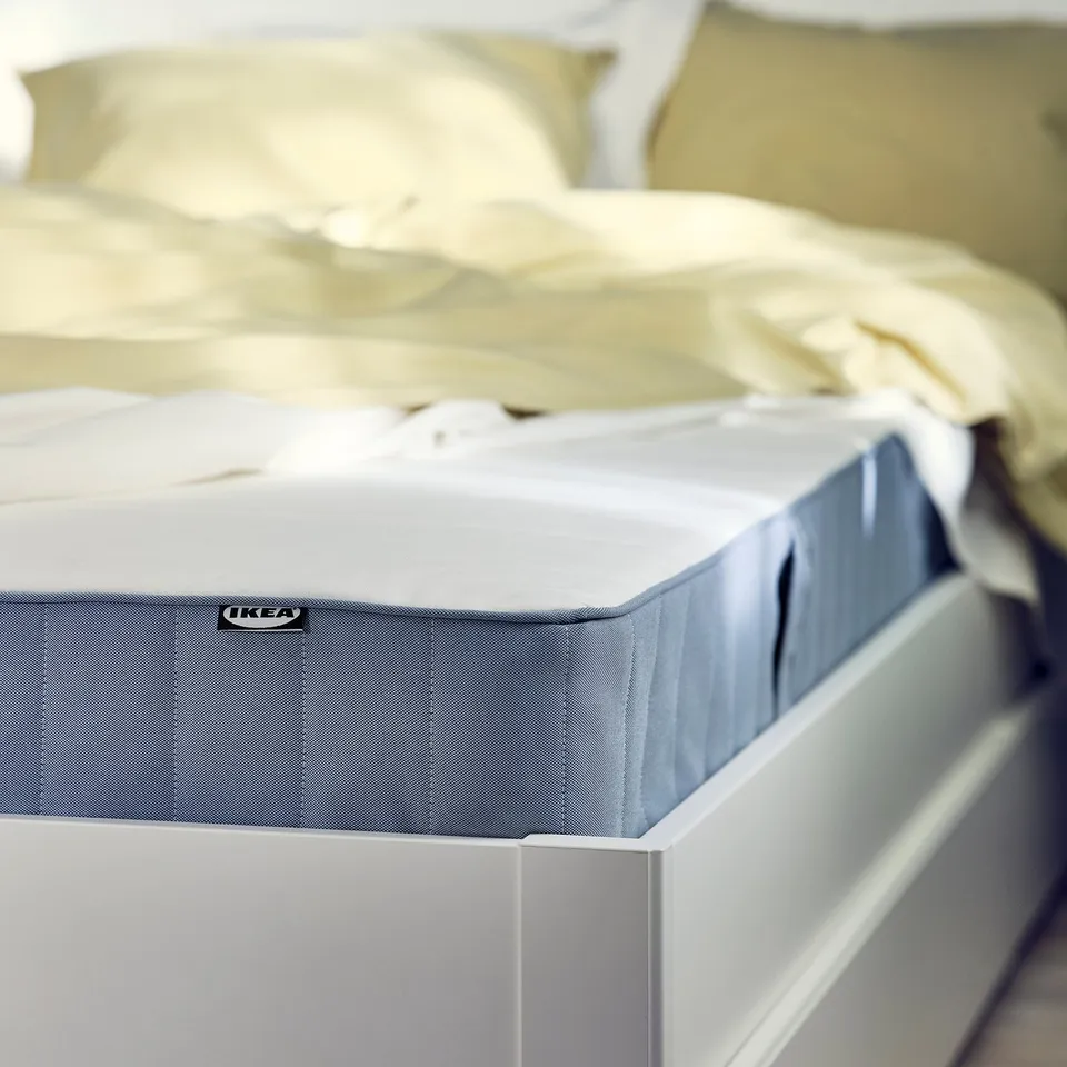 A close-up of a blue and white ikea mattress on a white bed, showing its firm construction with visible springs and quilted top.