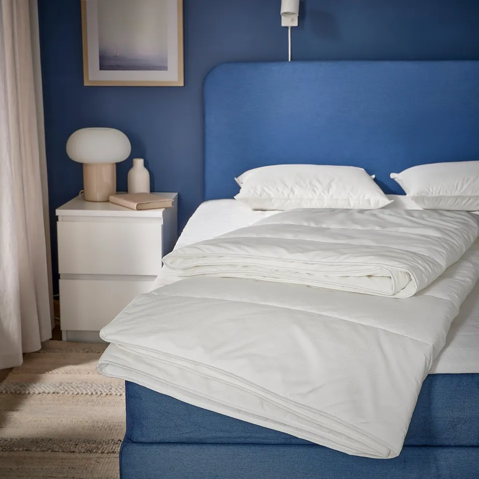 Cosy bedroom with SKOGSFRÄKEN duvet on blue bed. Fluffy white duvet, blue wall, white pillows, wooden bedside table with lamp and books.
