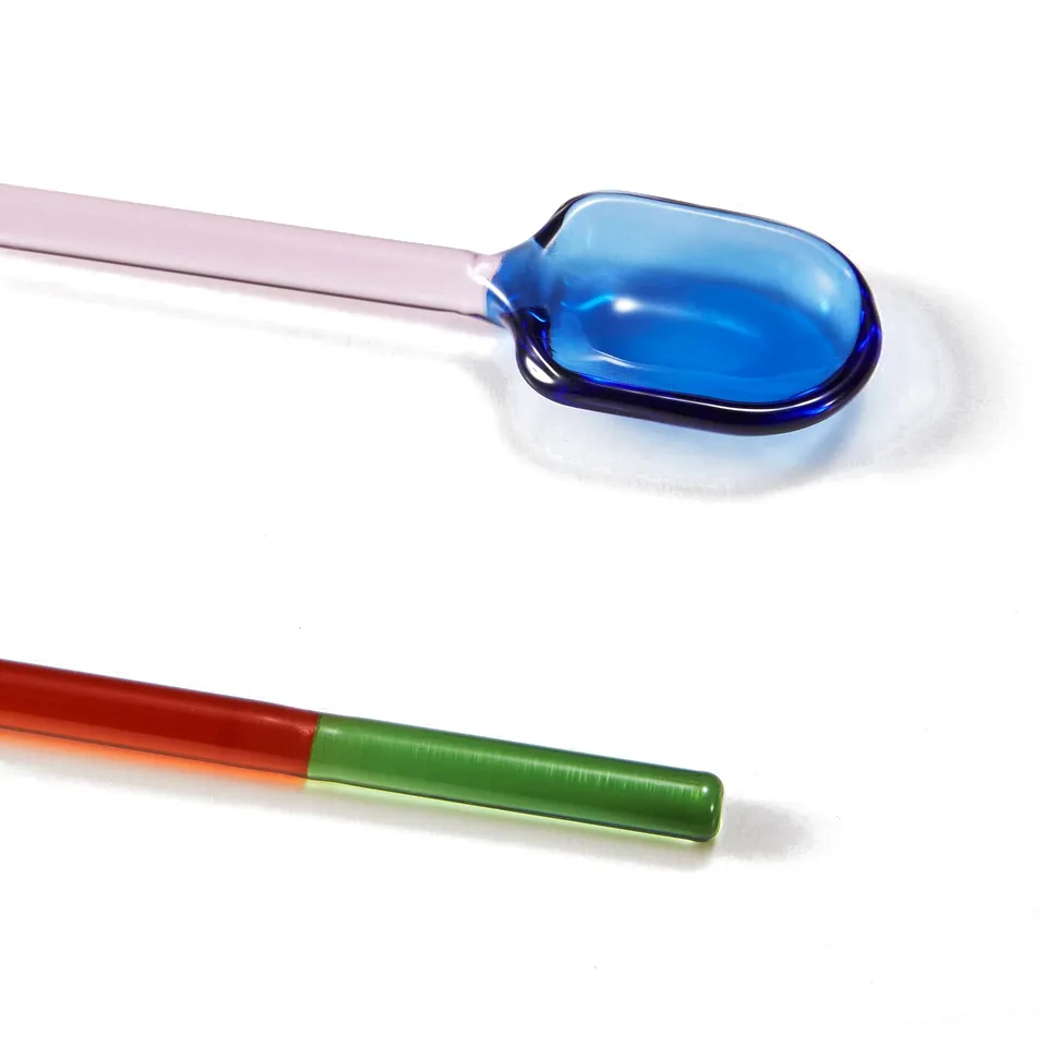 Two colourful glass spoons: one with blue and pink hues, the other with red and green sections, lying on a white surface.