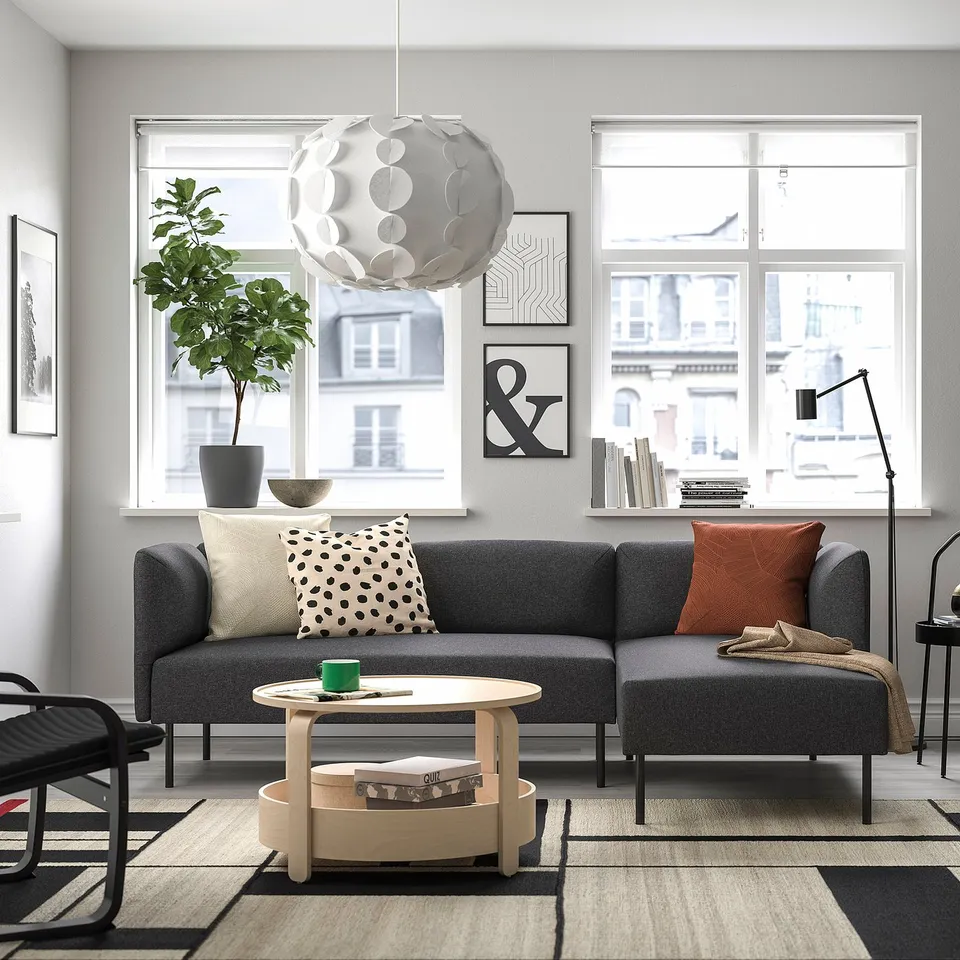 Modern living room: grey LILLEHEM sofa with cushions, wooden coffee table, large window, black chairs, green plant, and unique light fixture.