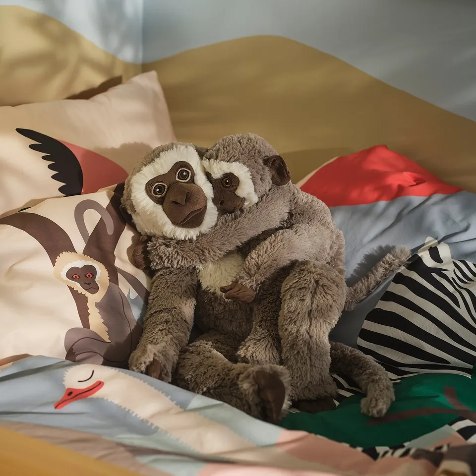 Two plush brown monkeys sit on colourful bedding, larger than nearby printed pillows.