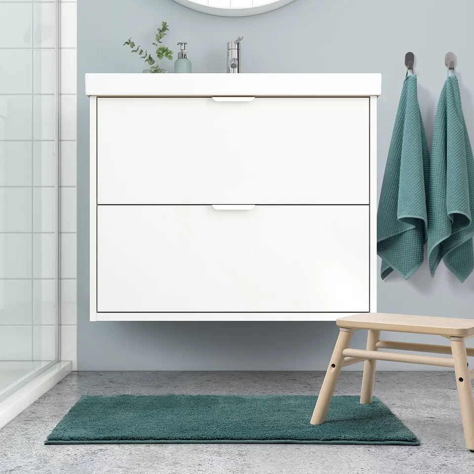 A modern bathroom with a white BACKMÅRA bath mat in teal, rectangular, soft, placed on a grey floor beside a white cabinet and wood stool.