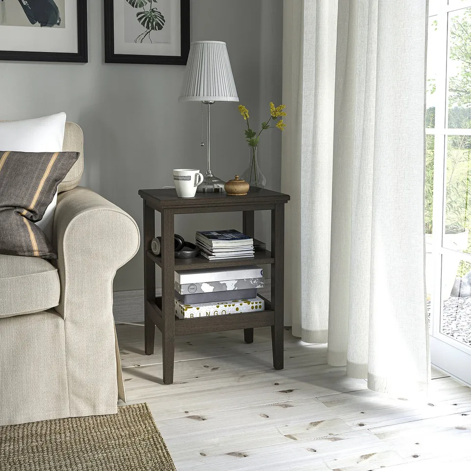 Wooden side table IDANÄS with shelves, beside armchair, grey & natural wood, near window with curtains.