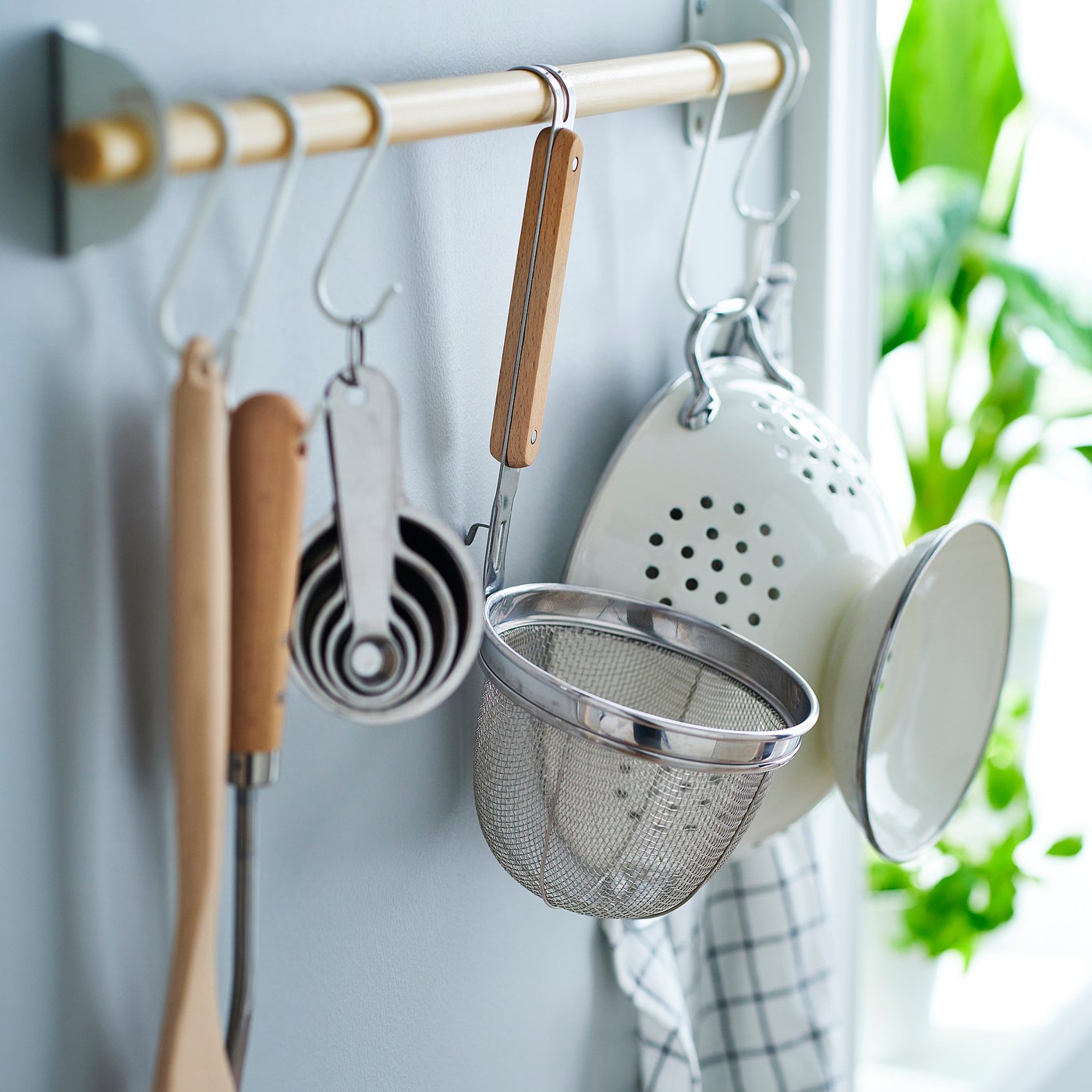 Metal colander with wooden handle hangs on kitchen rail, showcasing its deep basket for straining food.