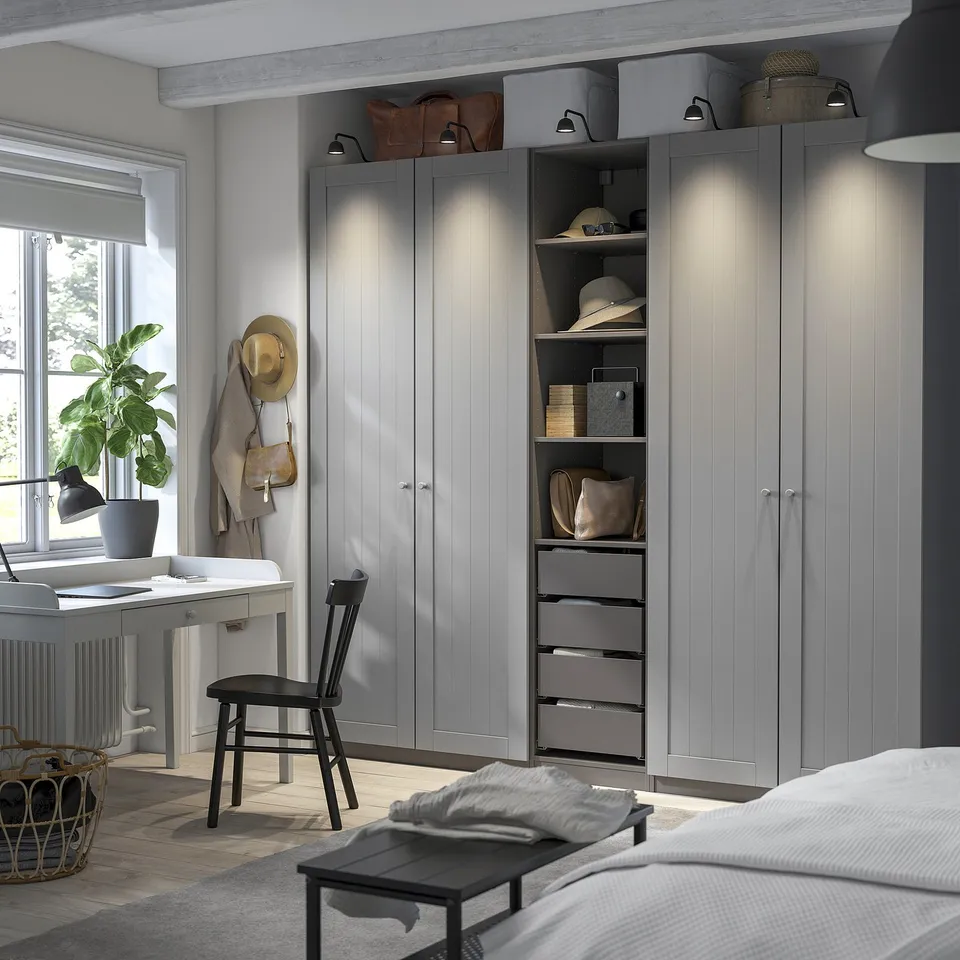 Modern bedroom with grey PAX wardrobe, integrated lighting, and storage. Beside bed: lamp, plant, hats, bags.