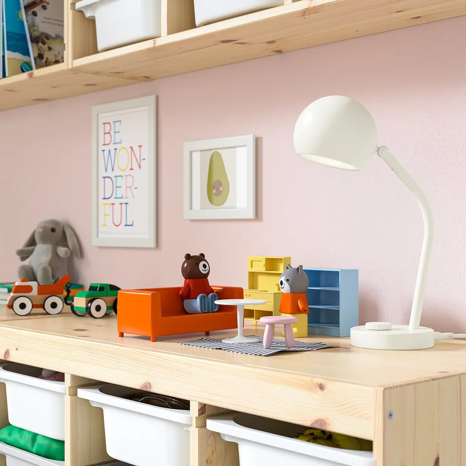 A childs room a wooden table with a white KORVSNÖRE lamp, smaller than the toy figures, shining on various colourful toys and a cosy teddy bear.