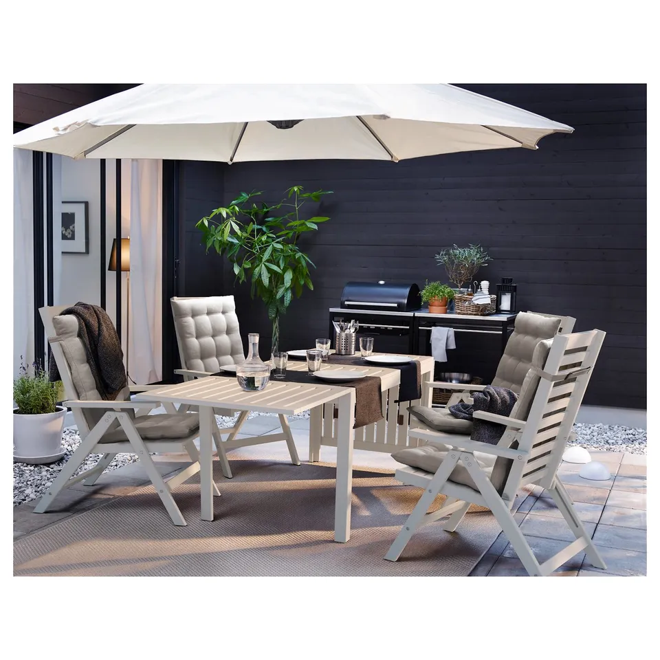 Outdoor patio set with white chairs, table, and rug under black wall. Large beige umbrella provides shade.