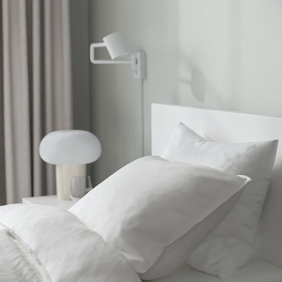 Neat bed with white bedding, lamp, and wall light, serene atmosphere.