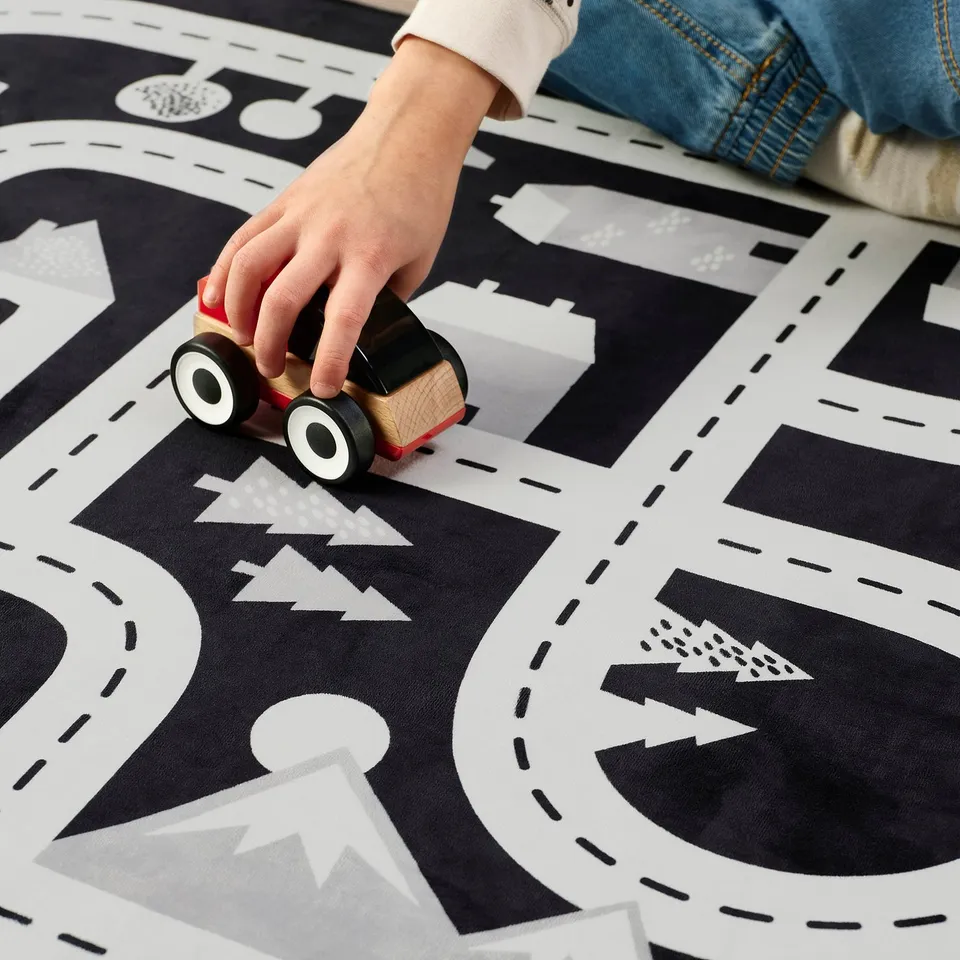 Childs hand moves wooden car on black and white play rug with roads and buildings, demonstrating interactive play.
