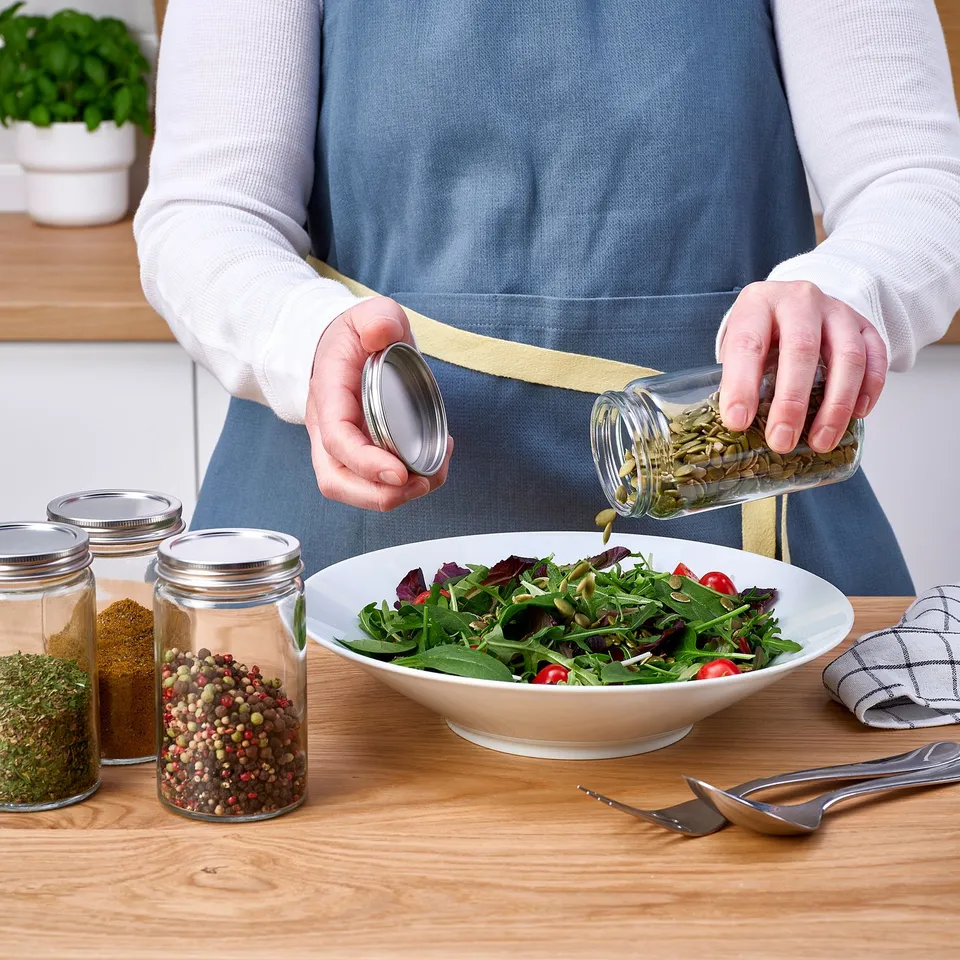 Person seasoning salad with herbs from a glass jar on a wooden table. Other spice jars are nearby.
