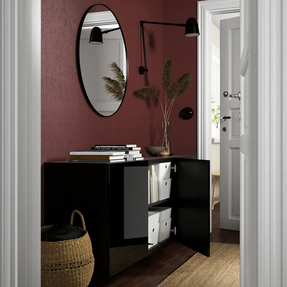 Black BESTÅ cabinet with open doors revealing adjustable shelves. A mirror, light, plant, and books sit on top.