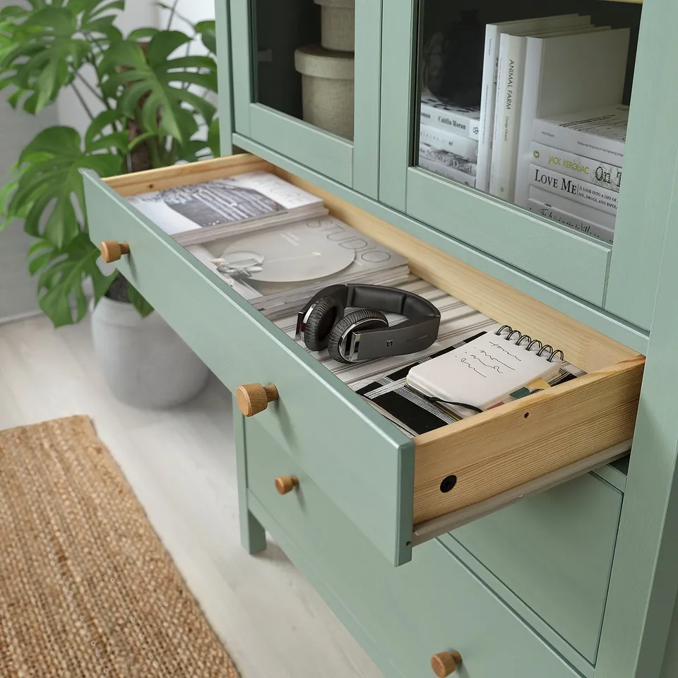 Green cabinet with open drawer showing headphones and papers, with books and plant nearby.