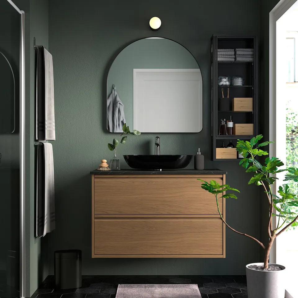 Modern green bathroom with large mirror, wooden vanity, black sink, plants, grey towels.