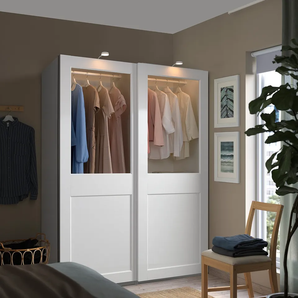 A PAX wardrobe with white sliding doors, displaying colourful clothes. It stands tall, surrounded by smaller furniture.