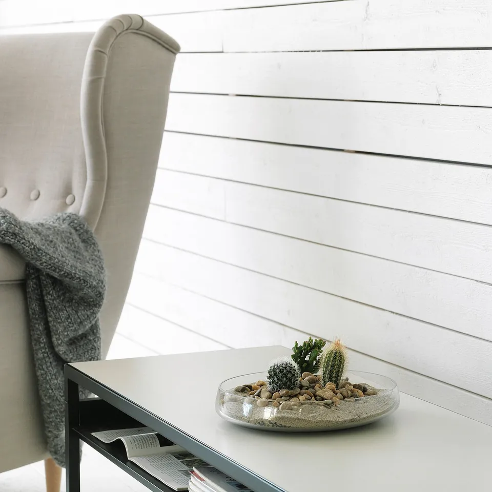 Modern chair with beige fabric and grey blanket, glass bowl with small plants and stones on white table against white wooden wall.