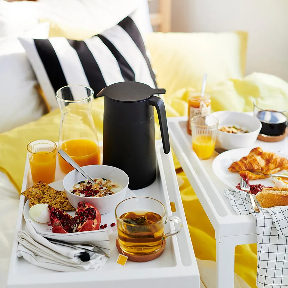 Bright breakfast trays on a bed, featuring juice, tea, and pastries. Modern black coffee flask, white dishes, and yellow accents.