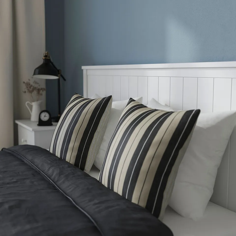 Cosy bed with GULLABERG striped pillows on dark blue bedding, white headboard, and bedside table with lamp and clock.