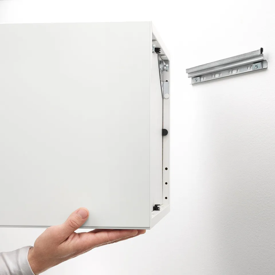 Person holding white EKET cabinet, showing wall mounting hardware.