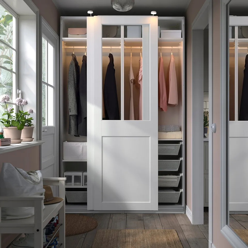 Modern white PAX wardrobe with glass/wood panels, sliding doors, near window and chair.