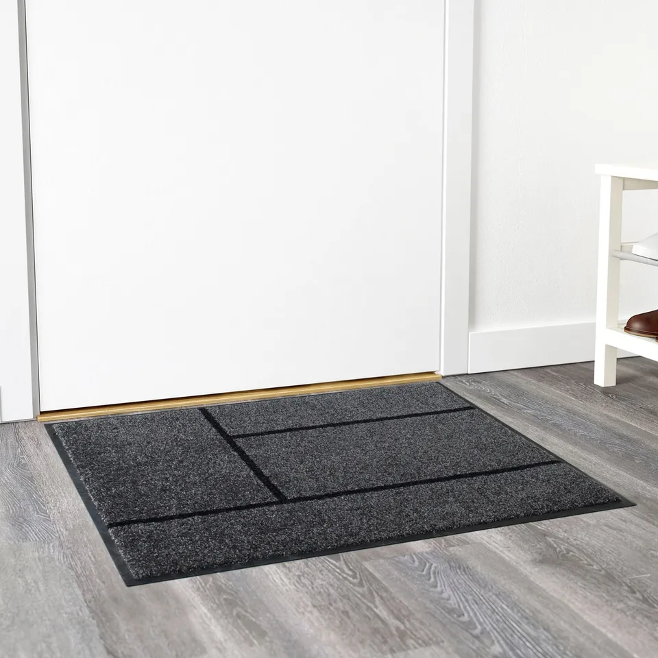Such as: dark grey door mat with black lines, lying flat on wooden floor, keeps entryway clean.