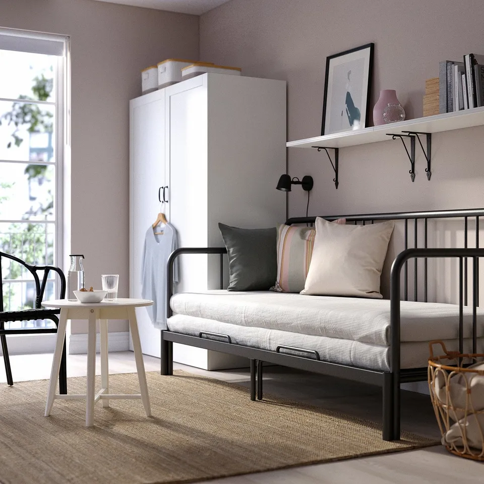 Modern minimalist bedroom with FYRESDAL grey day-bed, white table, grey rug, tall white cabinet, and wall shelf with books and vase.