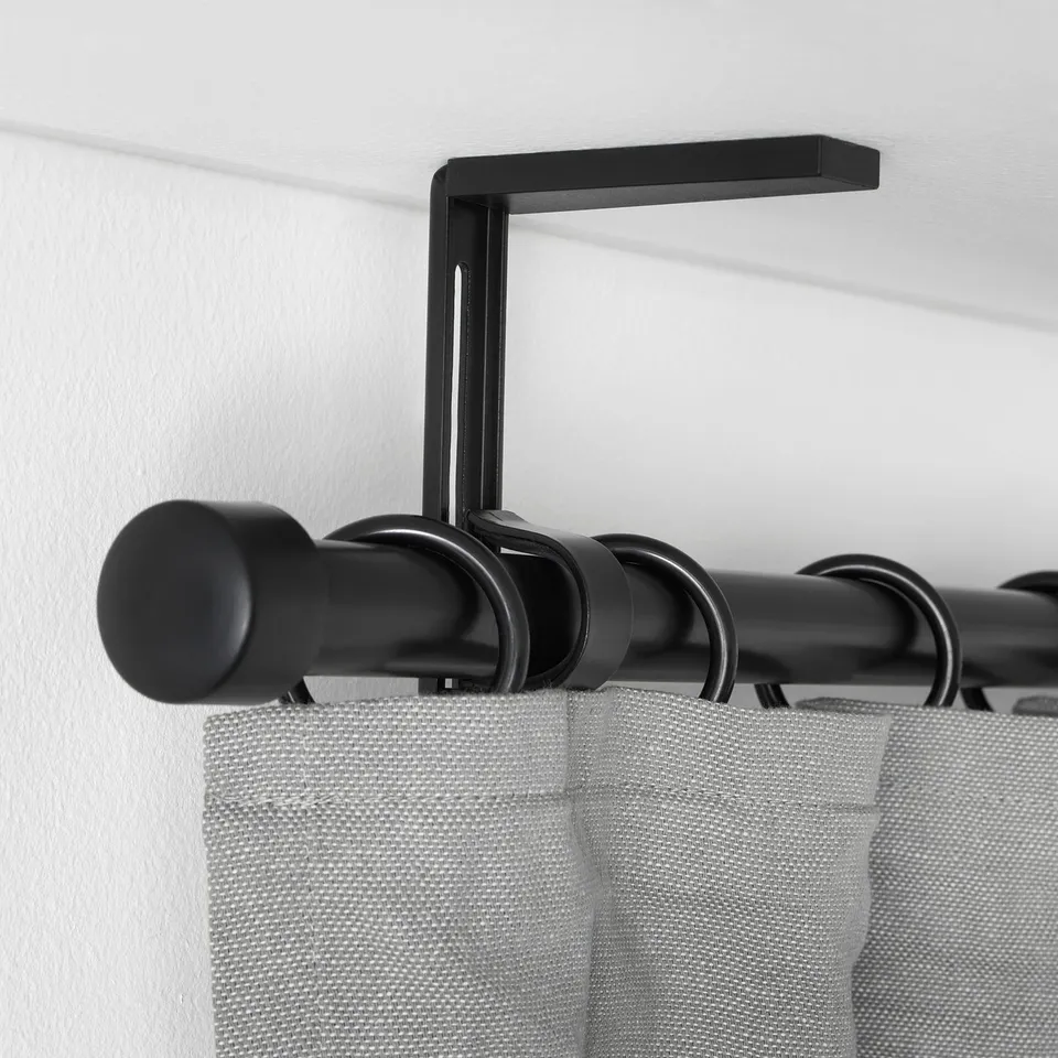 Black curtain rod holder mounted on white wall, holding black rod with grey curtains, demonstrating adjustable positioning.