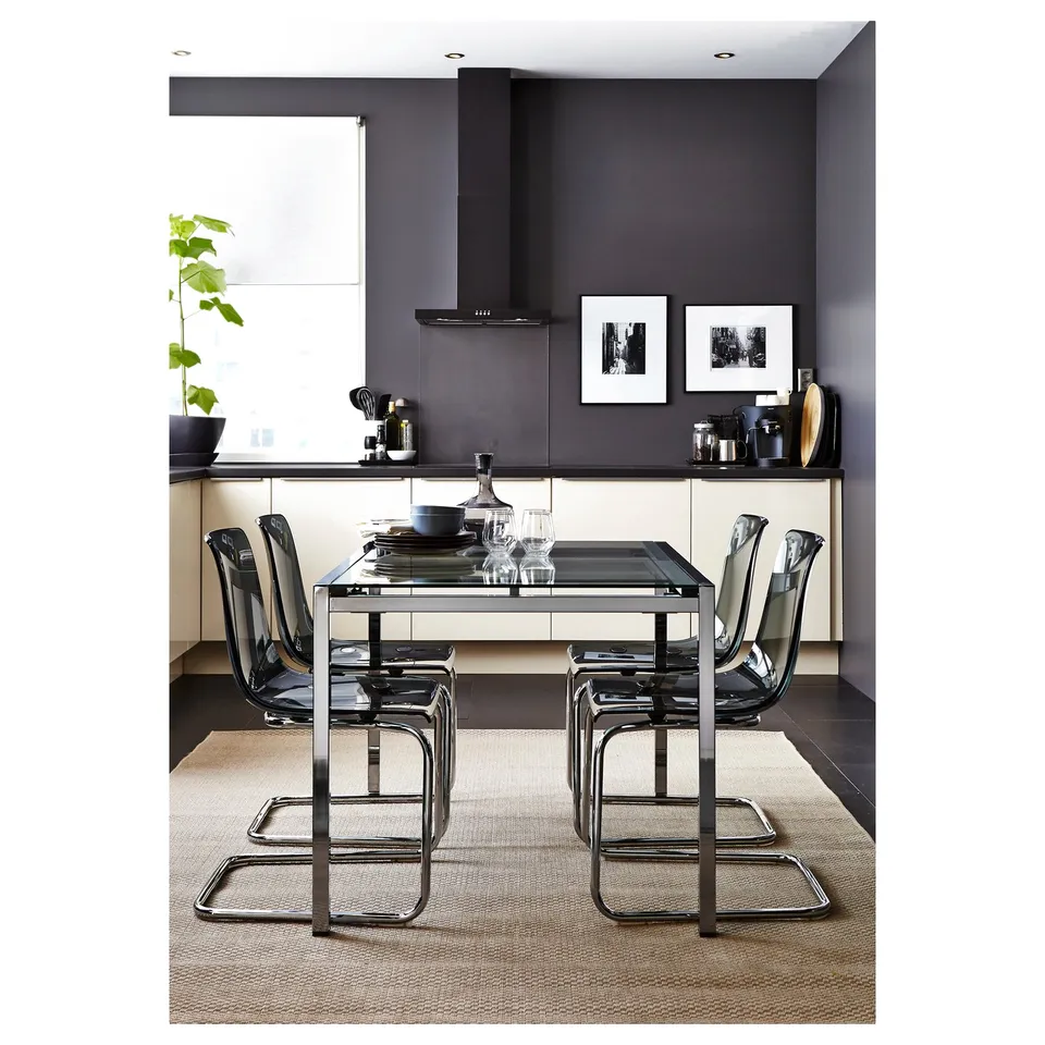Modern dining area with black glass table, four clear chairs, dark walls, white cabinets, and a patterned beige rug.