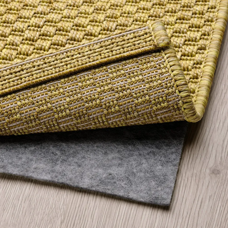 Green woven rug with white stripes, partially folded to reveal grey non-slip underlay underneath.