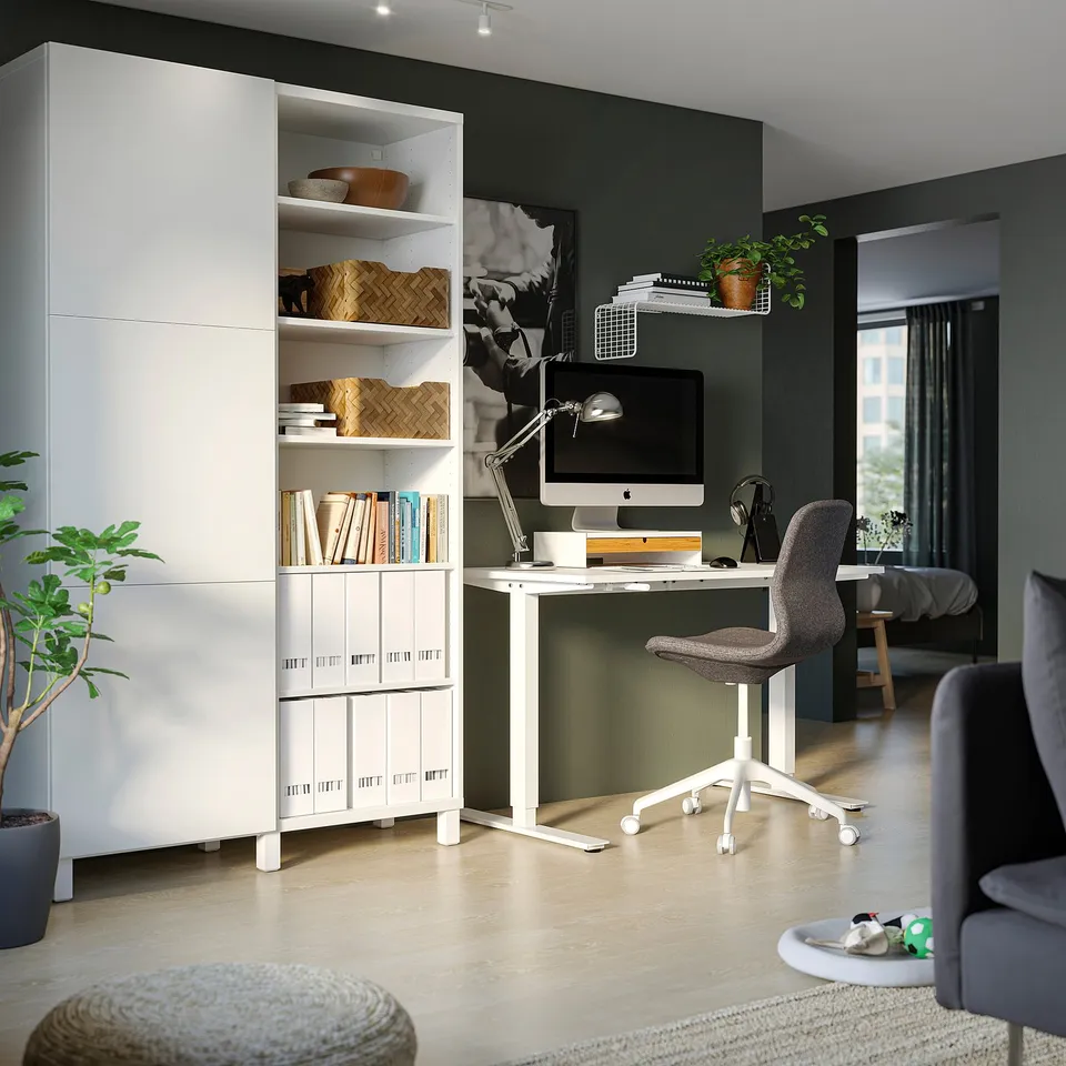 Modern home office with white BESTÅ shelving and desk, a grey chair, computer, and green plants.