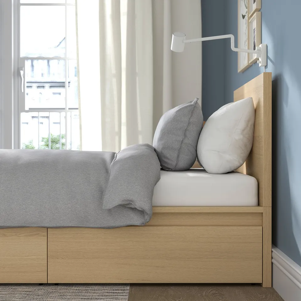 Modern wooden bed with grey bedding, two pillows, and built-in drawers. Wall-mounted lamp is attached to the headboard.