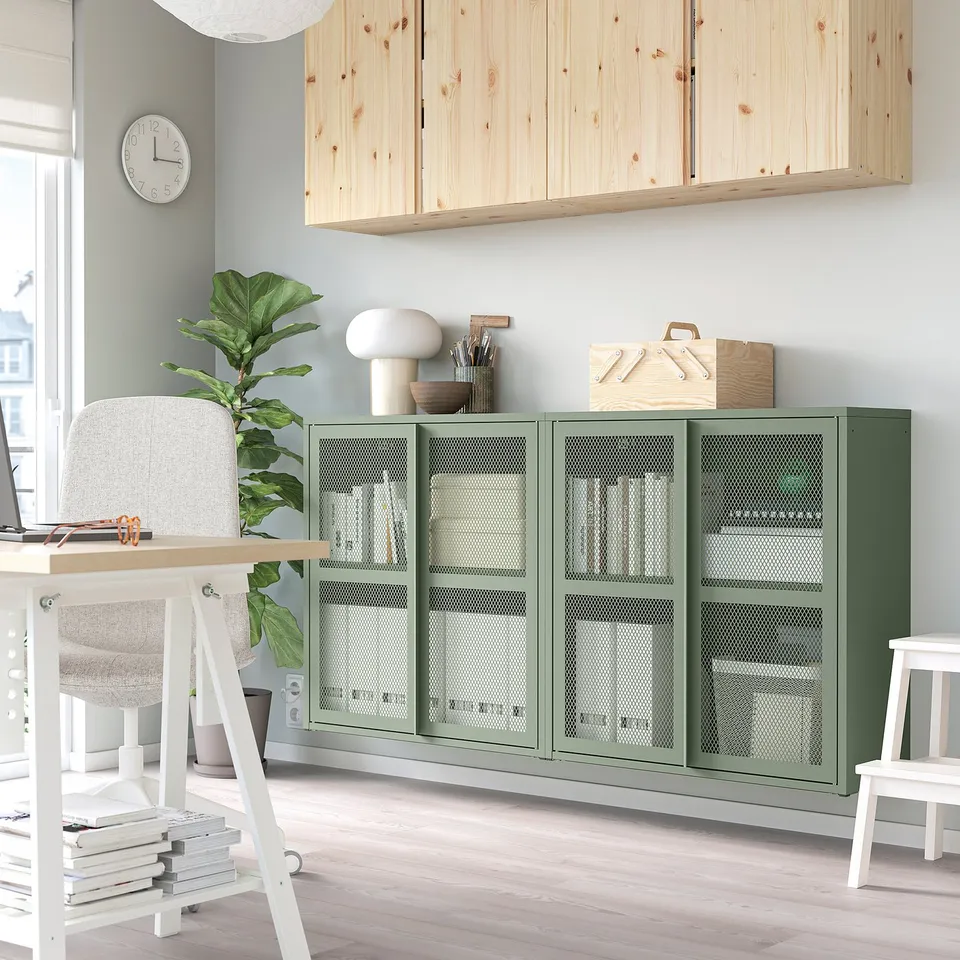 Modern room with IVAR green metal cabinets featuring mesh doors, large plant, white desk, and wooden shelves.