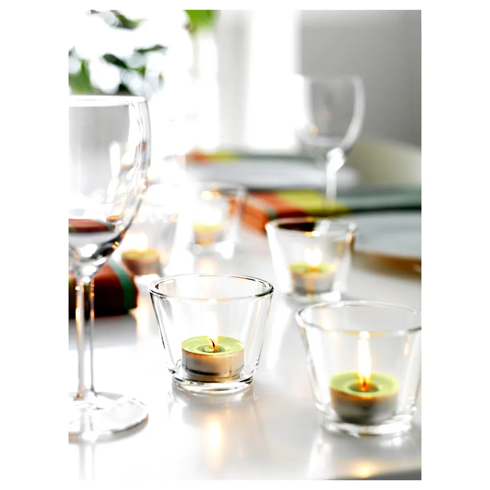 Three clear glass GALEJ tealight holders with lit candles on a table, surrounded by wine glasses.