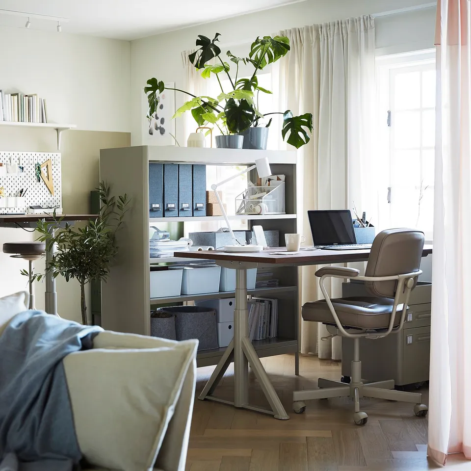 Modern home office with adjustable desk, laptop, chair, plants, and large window.