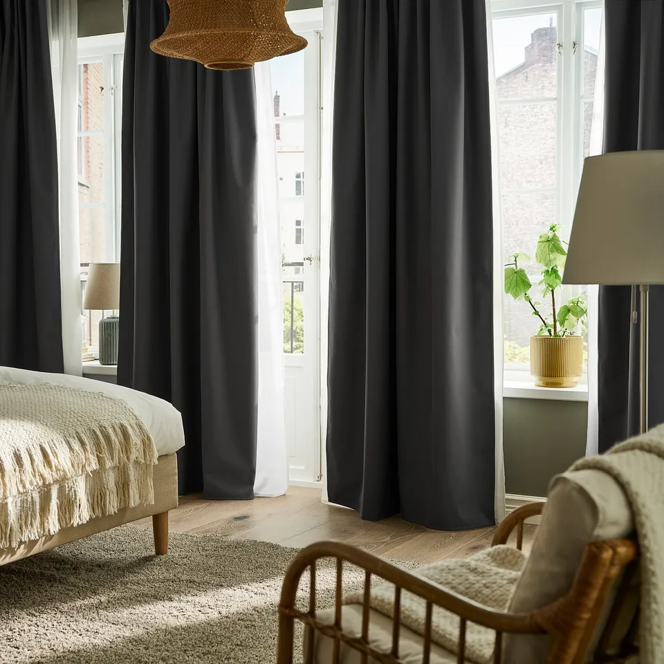 Minimalist bedroom with KORGMOTT dark grey curtains, wooden floor, white bed, wicker chair, and open balcony.