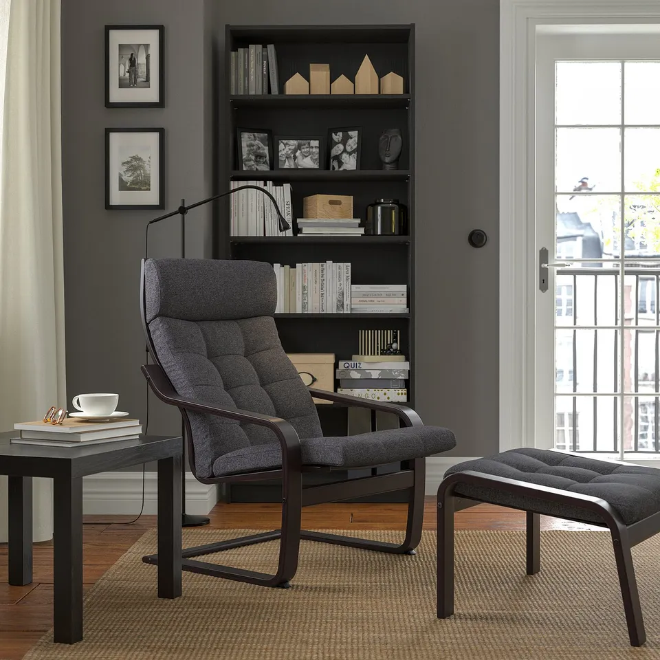 Grey POÄNG armchair with cushions, footstool, bookshelf, and window.