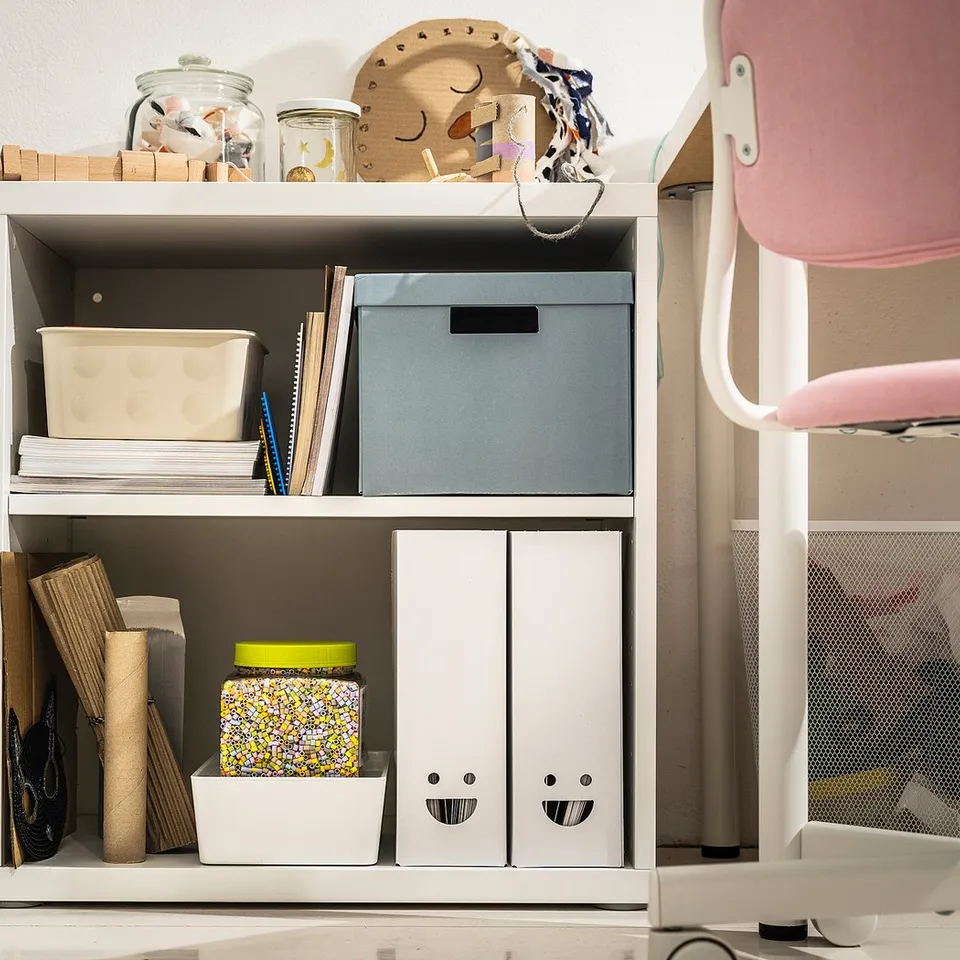 Organised bookshelf with colourful boxes and jars. Two white TJABBA boxes with smiley faces, blue and white bins, and a patterned bag.