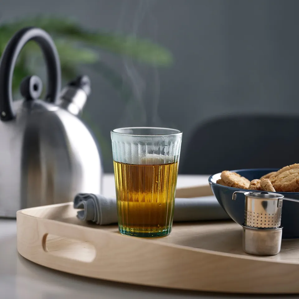 A wooden tray holds a glass of steaming tea, a bowl of cookies, and a tea strainer. Next to the tray is a silver kettle.