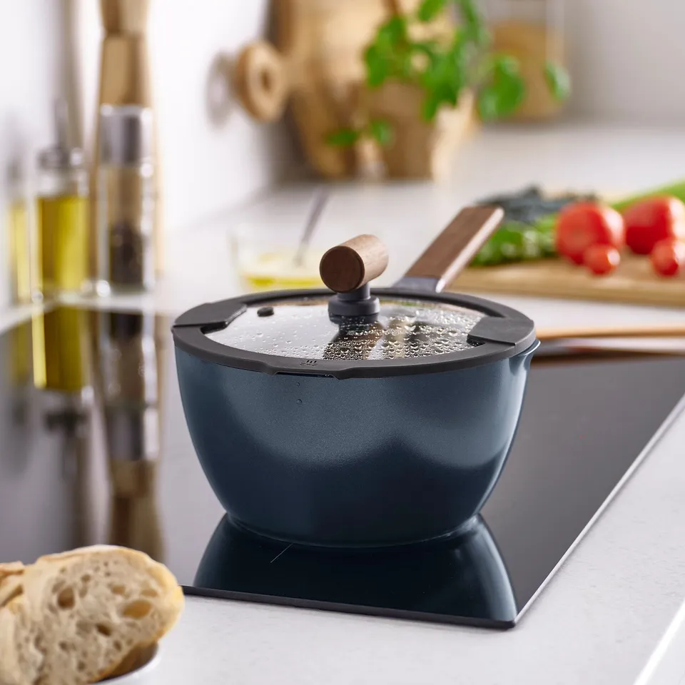 Black-blue cast aluminium saucepan with wooden handles and glass lid on kitchen counter.
