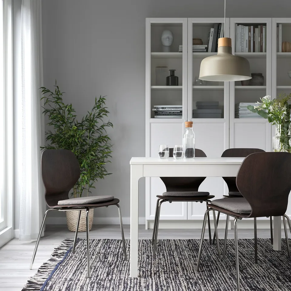 Modern dining set with white ÄLVGRÄSMAL chair pads, dark wooden chairs, and a rectangular table, surrounded by a rug, bookshelf, and plants.